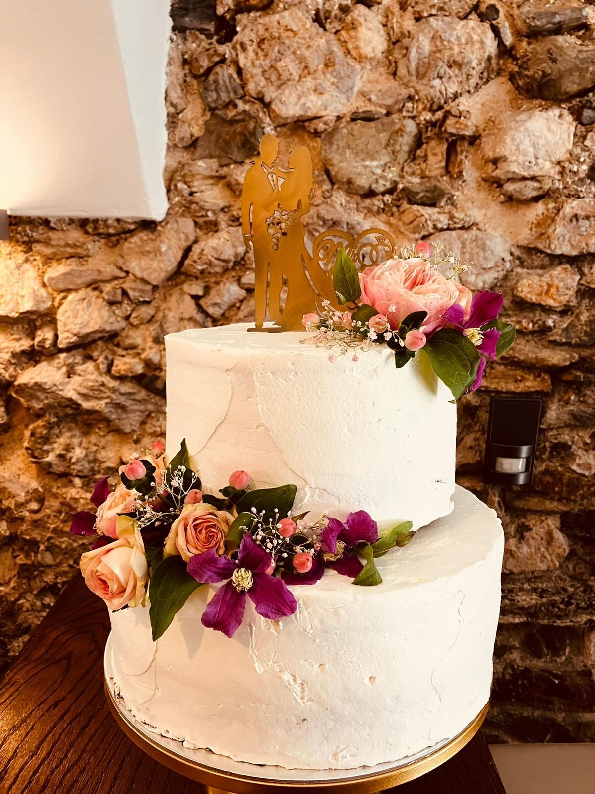 Eine Hochzeitstorte mit Blumen darauf steht auf einem Tisch vor einer Steinmauer.