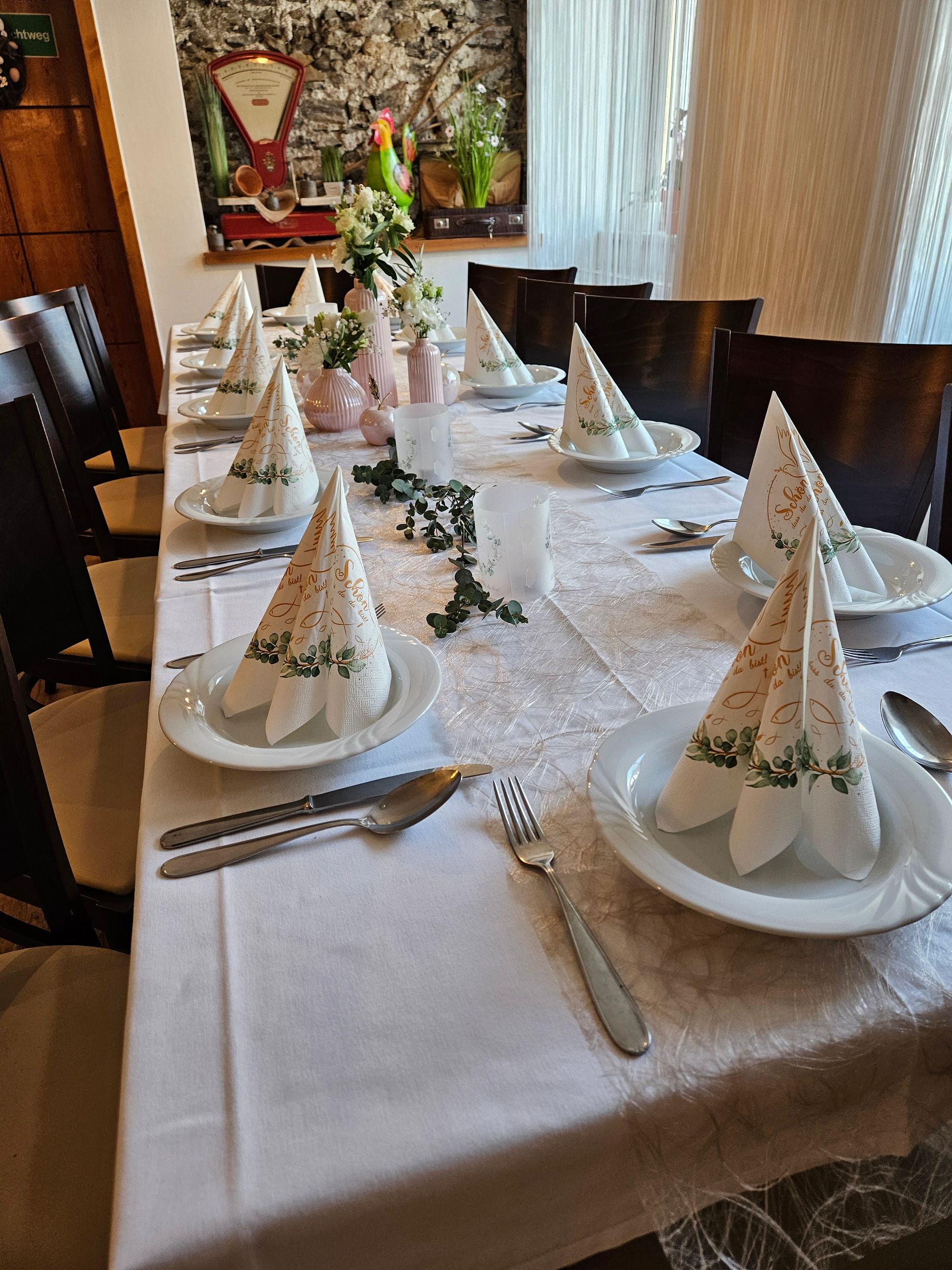 Ein langer Tisch mit Tellern, Servietten, Besteck und Blumen darauf.