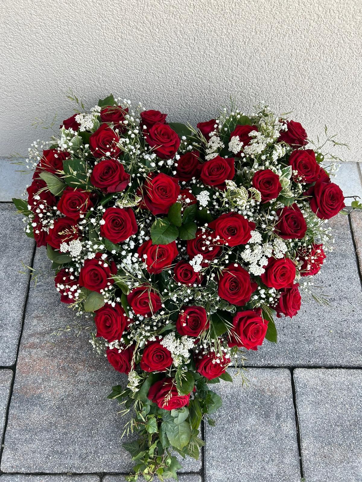 Auf einem gepflasterten Gehweg liegt ein herzförmiger Kranz aus roten Rosen und Schleierkraut.