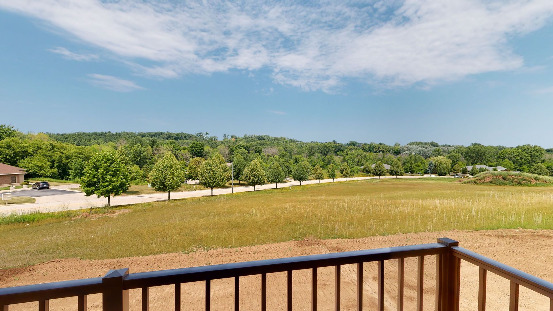 Wooden deck railing overlooking a grassy field, trees, and blue sky with scattered clouds