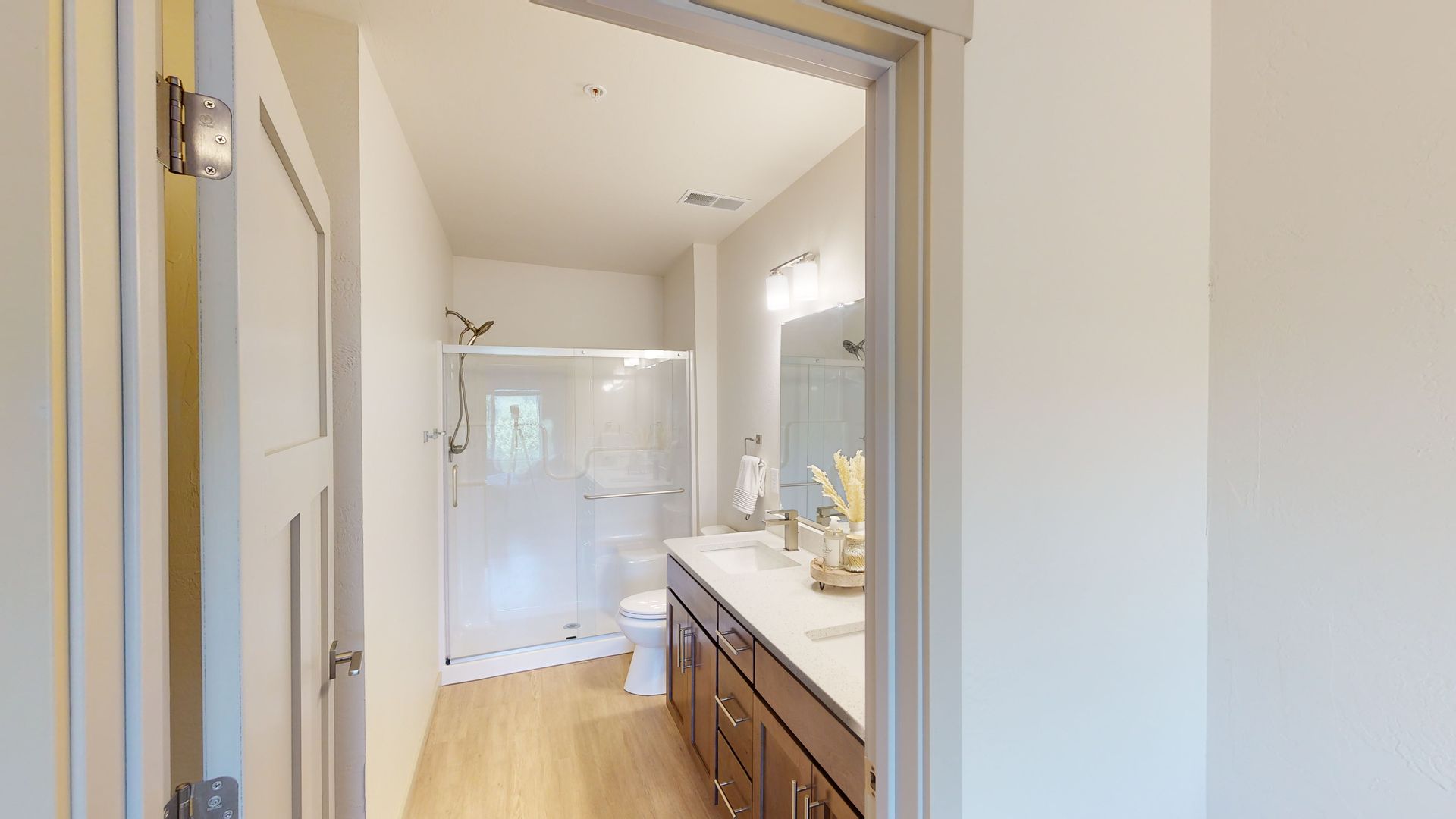 Bright bathroom with white shower, double vanity, mirror, and light wood flooring seen through open doorway