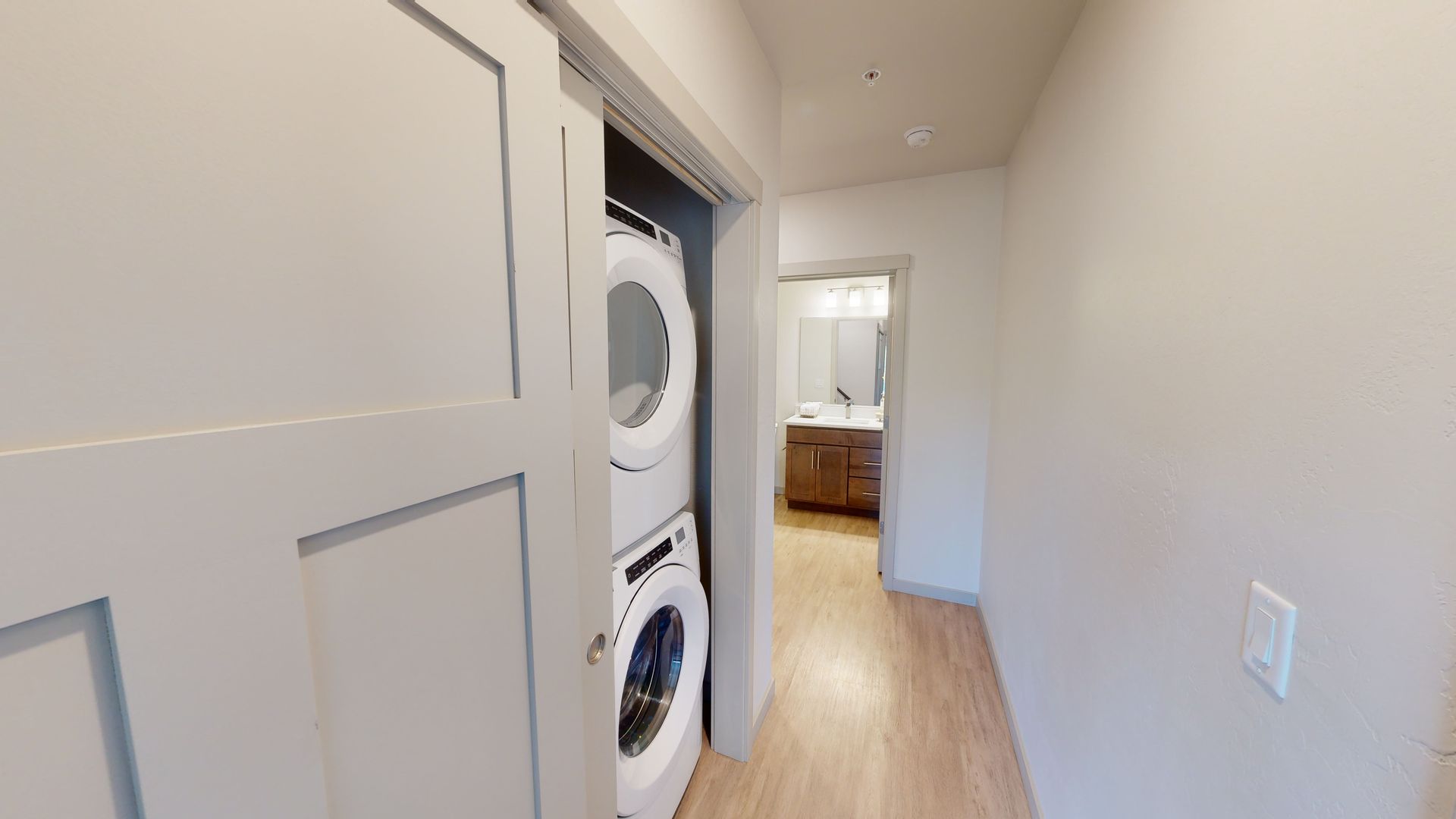 Narrow hallway with stacked washer and dryer in an open closet, light wood floor, and bedroom visible ahead