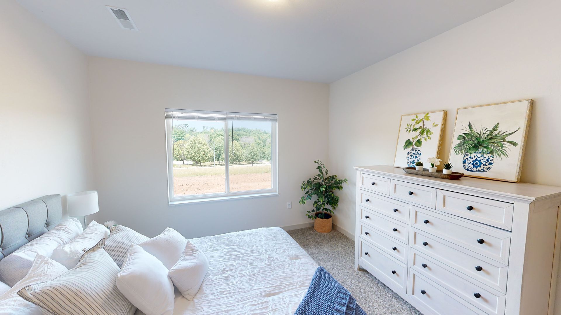 Bright bedroom with white bed, dresser, plant, and window letting in natural light