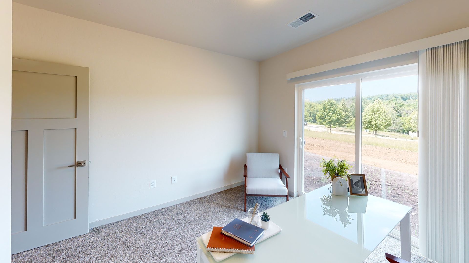 Bright empty living room with sliding glass door, beige walls, and white chair beside a glass coffee table.