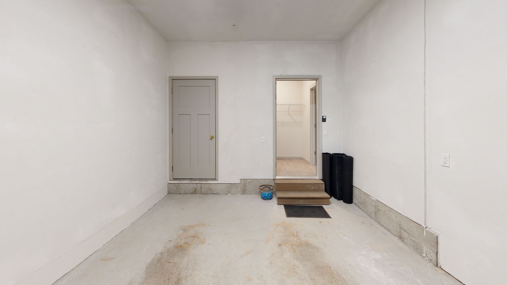 Empty white room with a closed door, open doorway, and dark floor mats near the entrance.