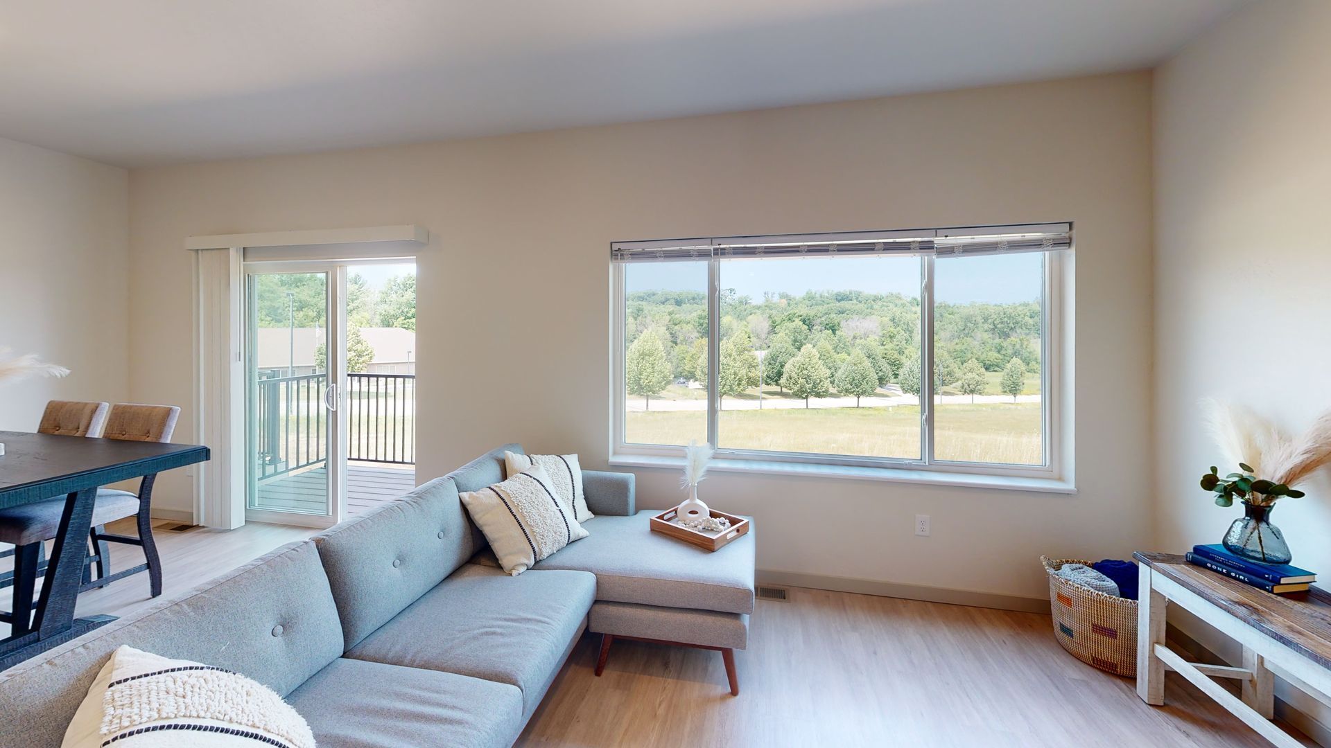 Bright living room with a gray sectional, large window, and a small table near a balcony door.