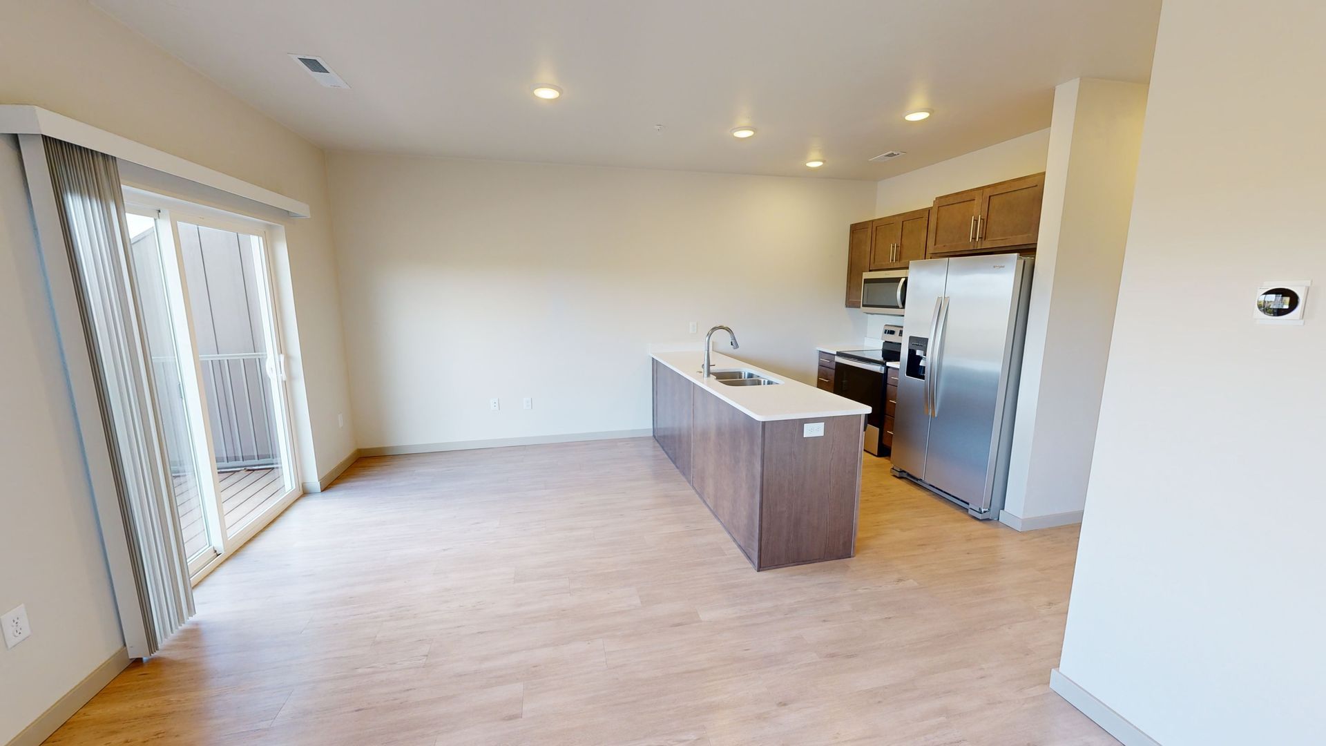 Bright open empty kitchen and living area with island, stainless appliances, light wood floors, and sliding glass doors.