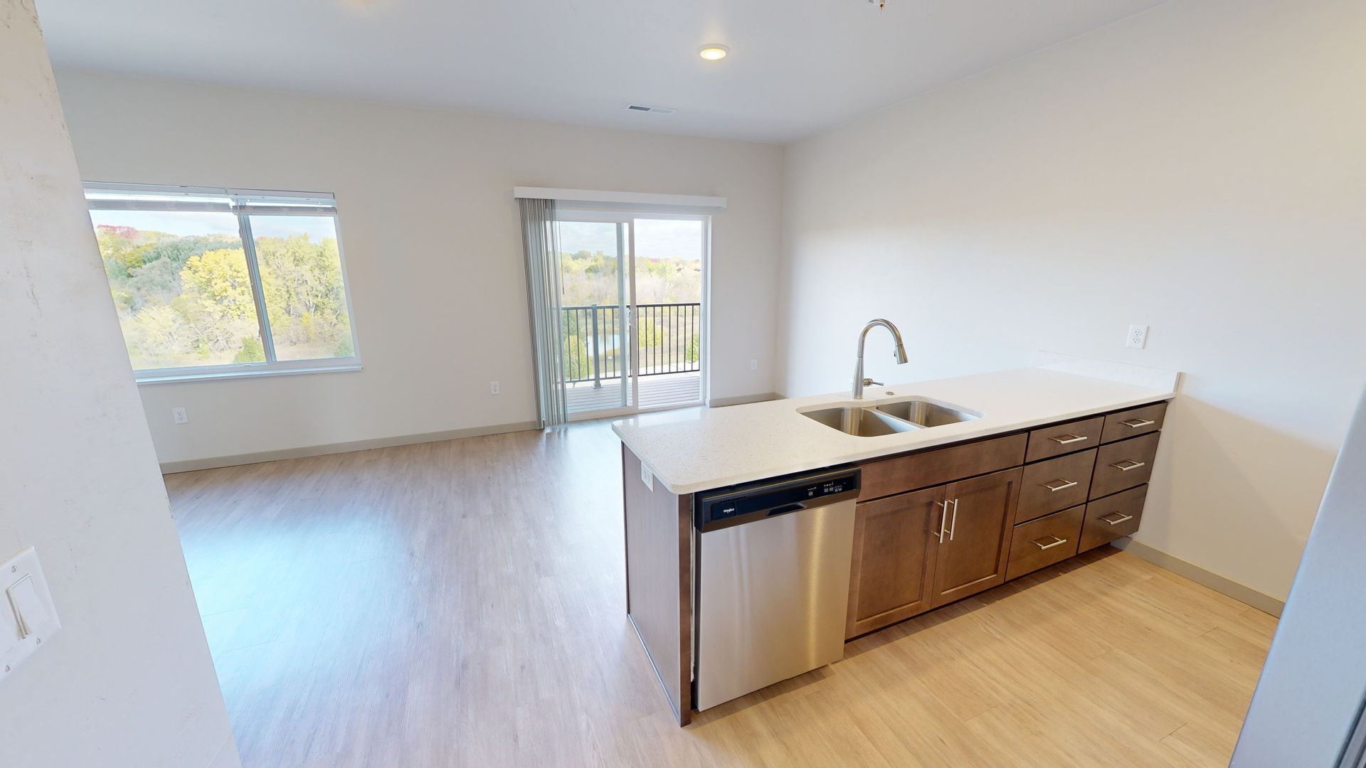 Empty modern kitchen and living area with light wood floors, white walls, and balcony doors.