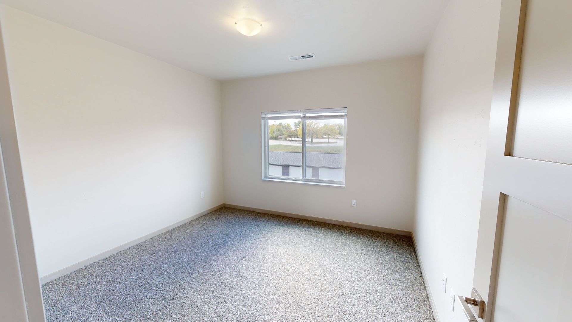 Empty bedroom with white walls, gray carpet, and a window letting in daylight.