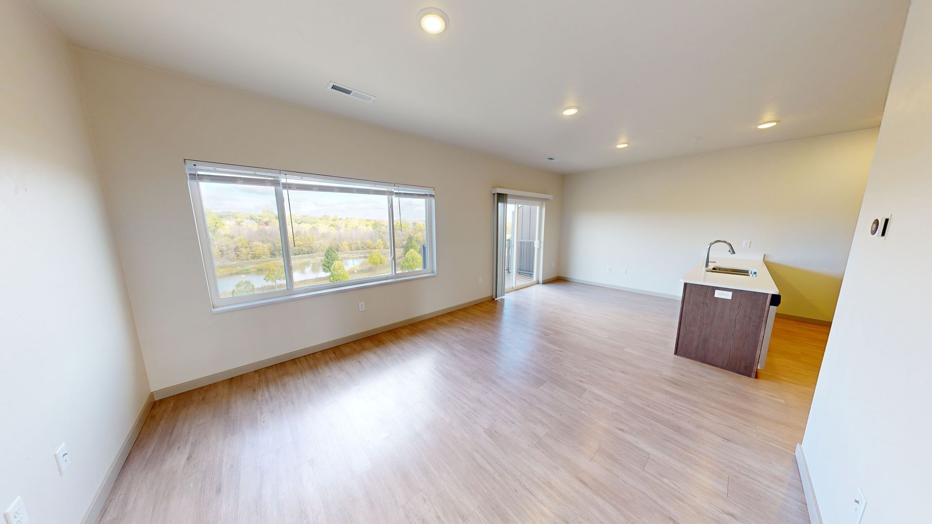 Spacious empty living room with wood floors, large window, recessed lights, and a kitchen island on the right