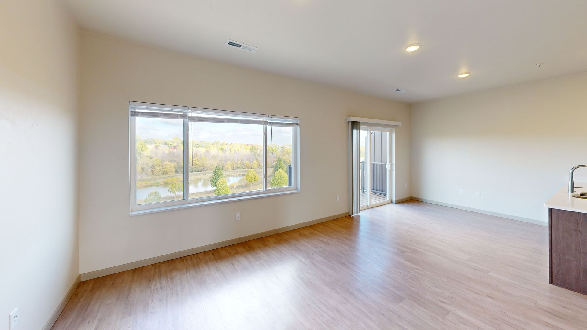 Empty modern living room with light wood floors, beige walls, and large windows.