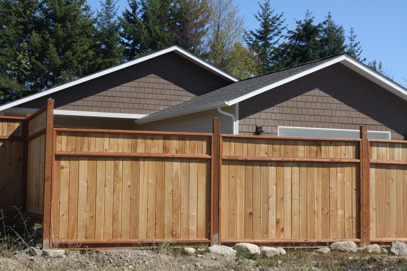 Wooden privacy fence in front of a house with a brown shingled roof, trees in the background.