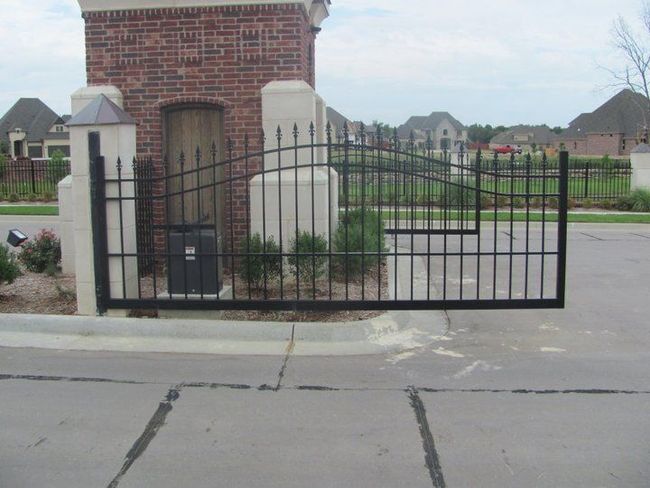 Black metal gate at the entrance to a gated community with brick pillars, a door, and houses in the background.