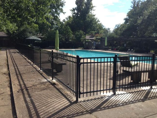 Black fence surrounds a swimming pool with lounge chairs and umbrellas. Sunlight casts shadows on the concrete.