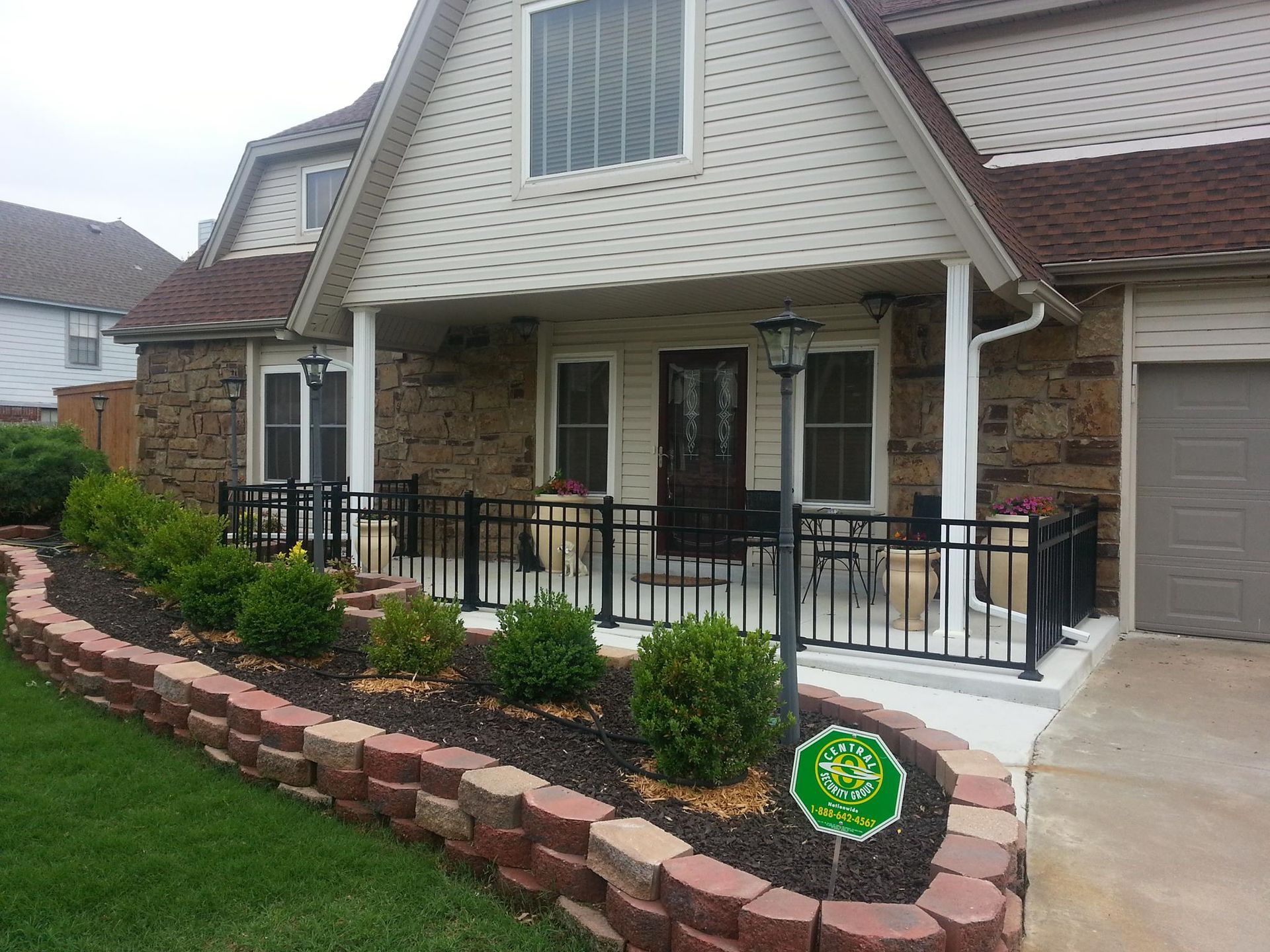 House with a small porch and black railing, stone siding, and a red brick flower bed with green bushes.