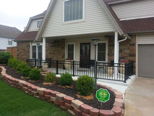 House with a porch, brick wall, flower beds, and black railing. Green grass and overcast sky.
