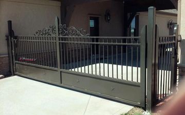 Brown metal gate blocking a driveway. Scrollwork top, vertical bars. Concrete surface, stucco wall background.