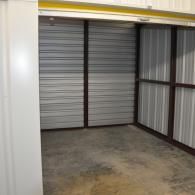 An empty, rectangular storage unit with corrugated metal walls, a concrete floor, and a metal roll-up door at the top.
