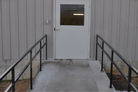 A concrete ramp with black metal handrails leading to a white door on a gray metal-sided building.