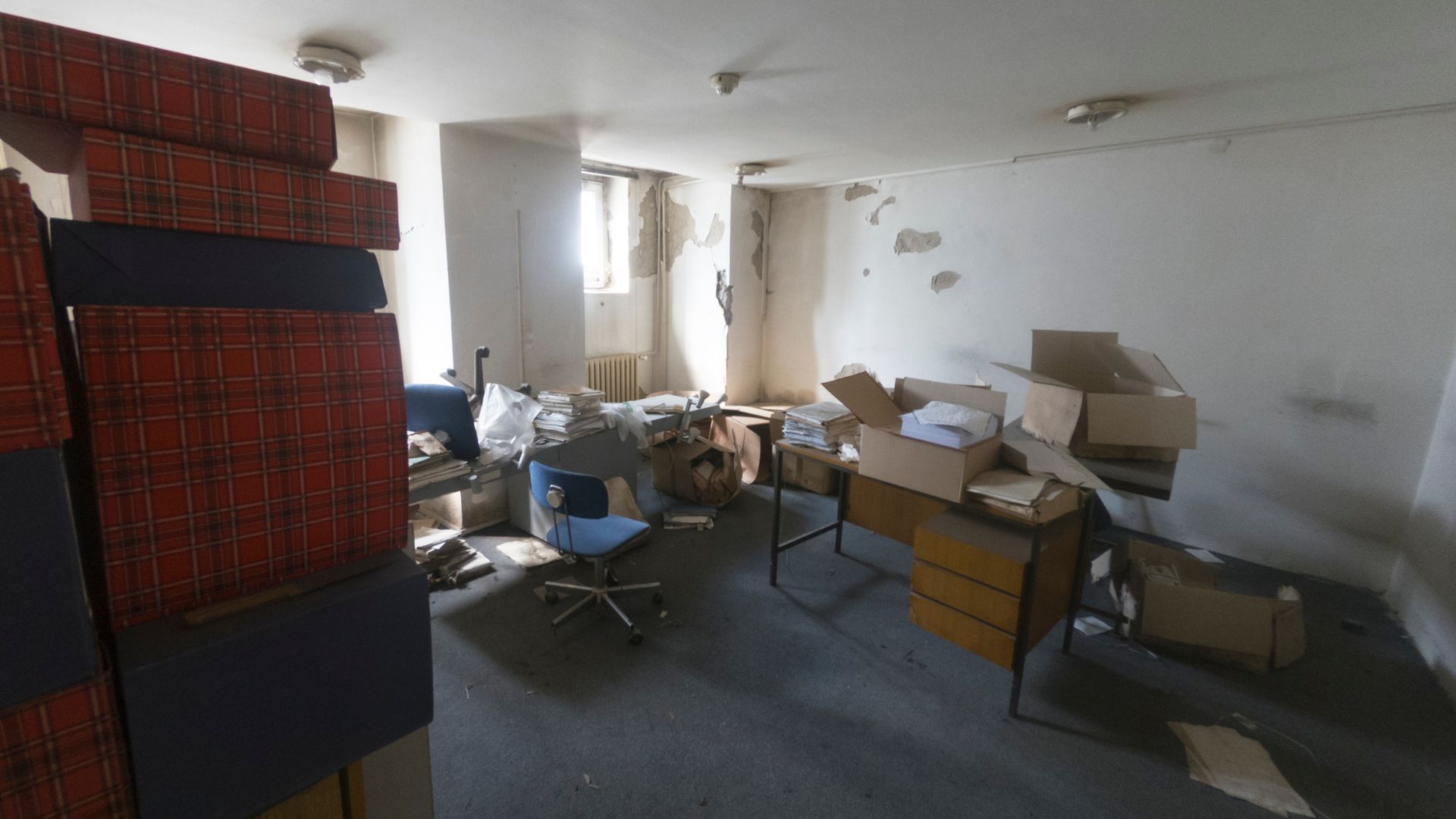A disused office with chipped paint on the walls, some wooden desks and chairs and cardboard boxes.
