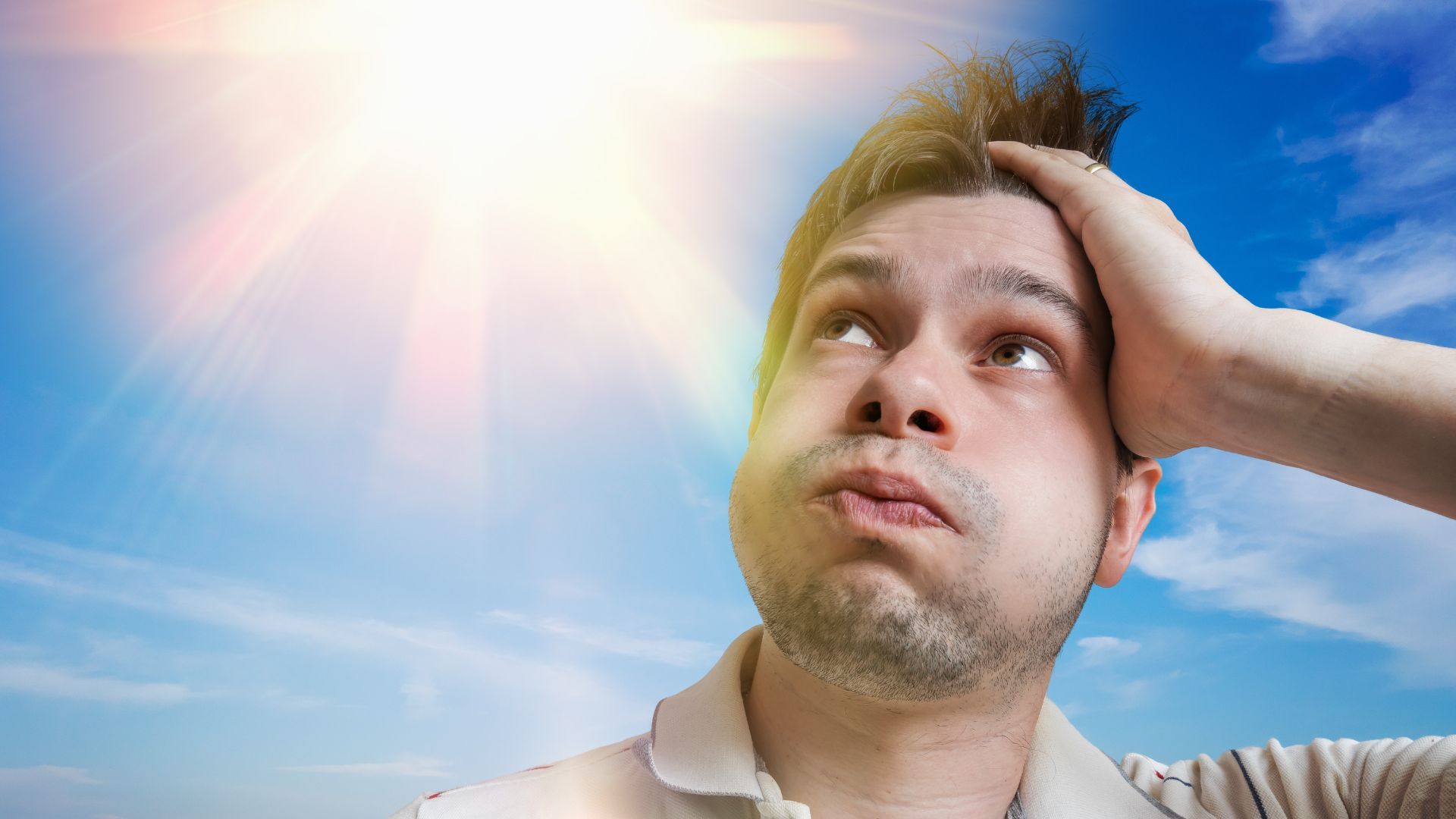 A man with his hand on his brow looking hot, sitting under the sun on a bright day with blue skies.