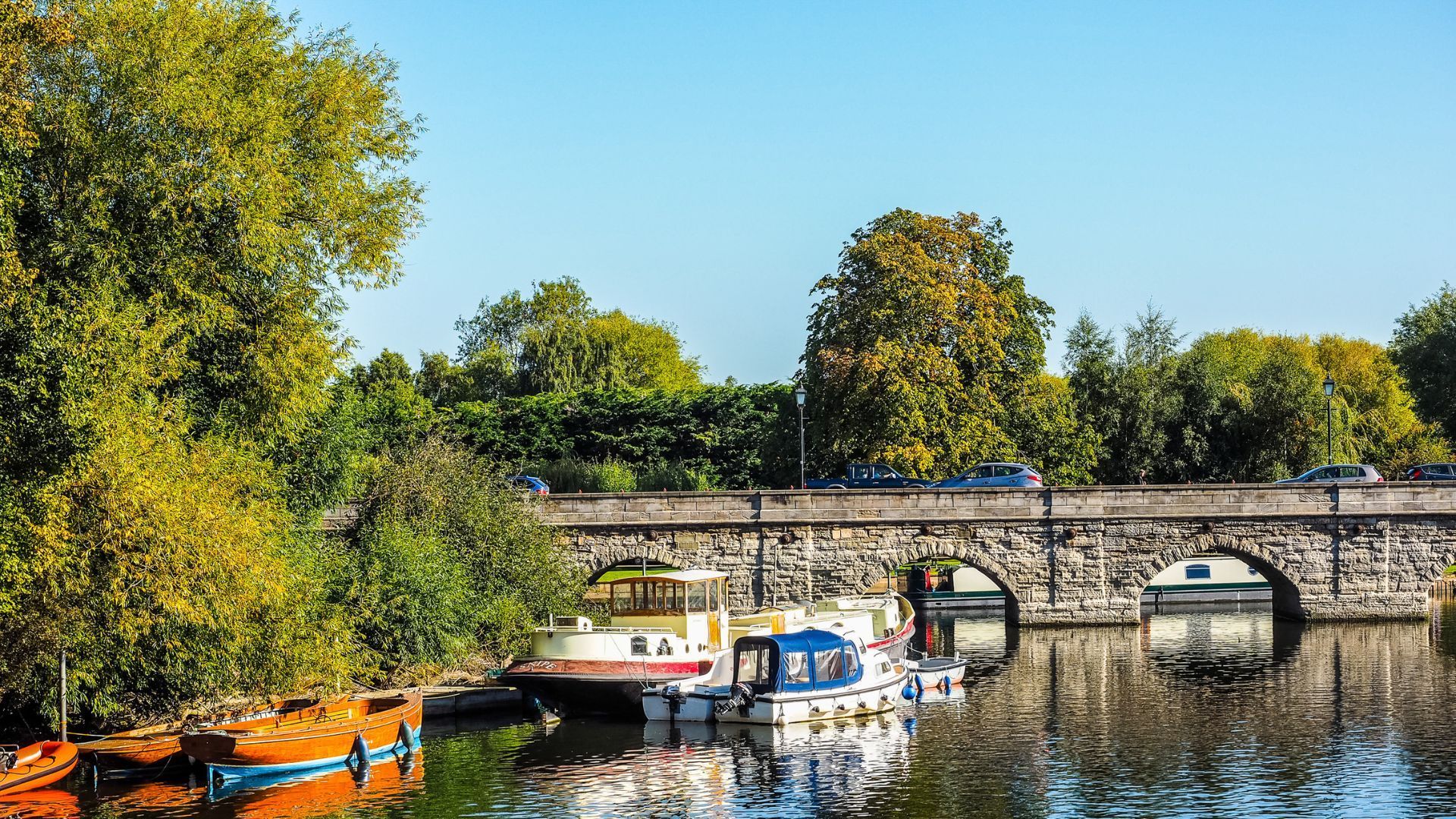 A scene of Stratford-upon-Avon showing the Tramway Bridge on a sunny day with blue skies, green trees and barges on the River Avon.