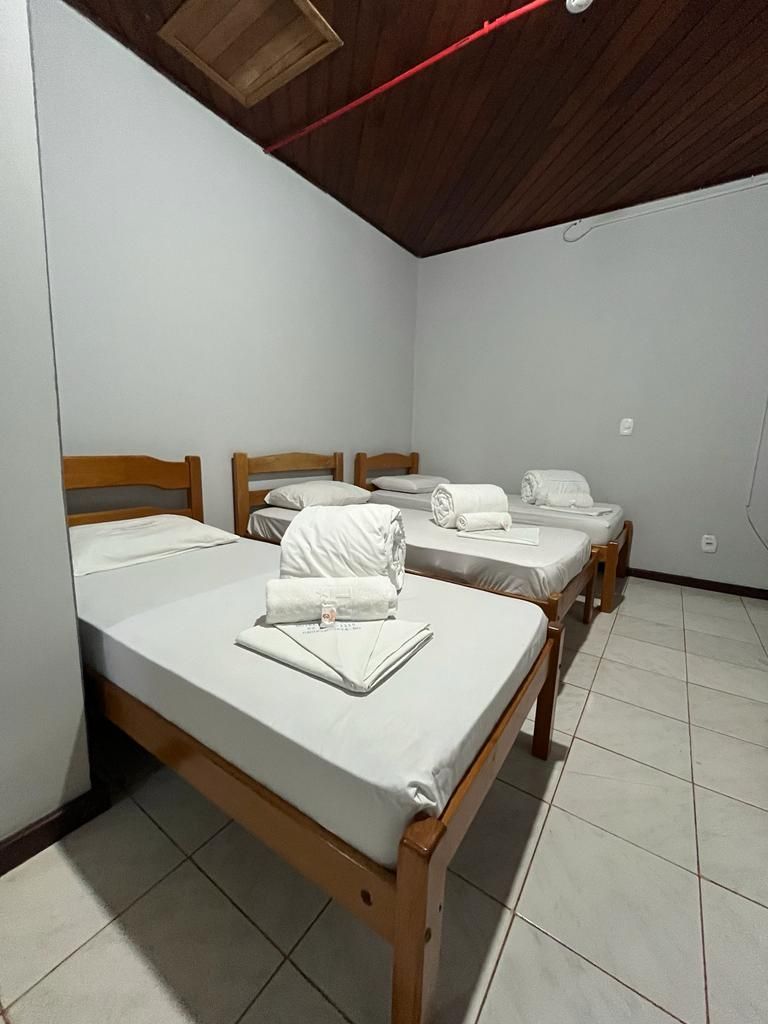 Quarto pequeno com três camas de solteiro de madeira, lençóis brancos e piso de azulejo sob um teto inclinado de madeira.