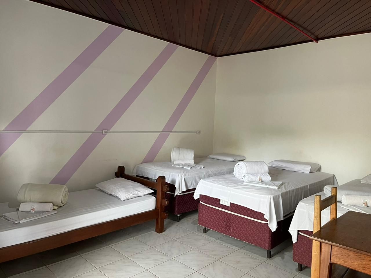 Quarto simples com quatro camas de solteiro, lençóis brancos e listras diagonais roxas na parede, sob um teto de madeira.