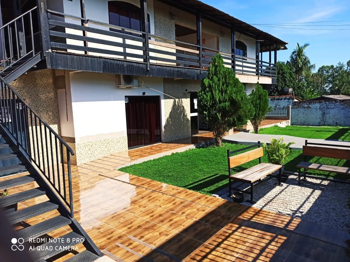 Pátio ensolarado com casa de dois andares, escada de madeira, varanda, gramado e terraço com bancos.