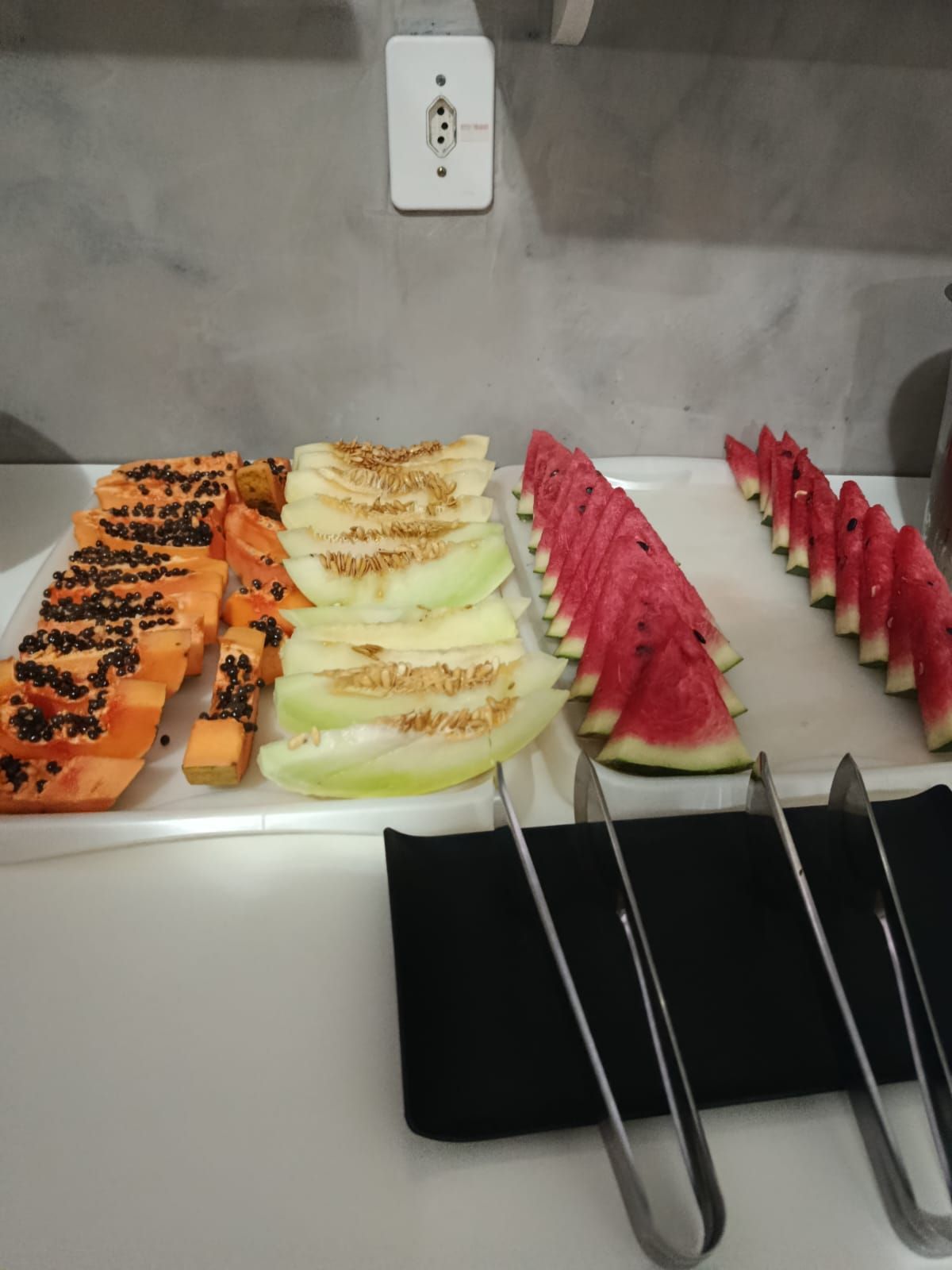 Bandejas com diversas frutas fatiadas sobre um balcão, incluindo mamão papaia, melão, melancia e pitaya.