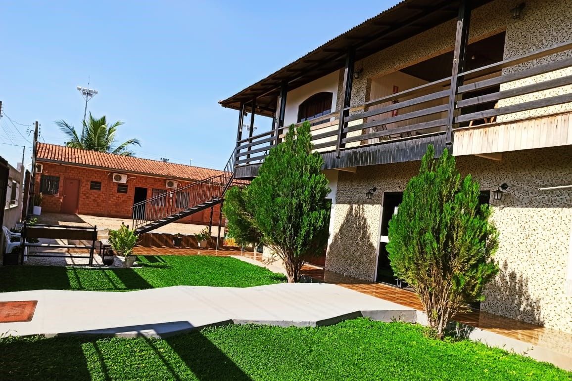 Pátio com gramado verde, calçada pavimentada e um prédio de dois andares com varandas de madeira sob um céu azul.