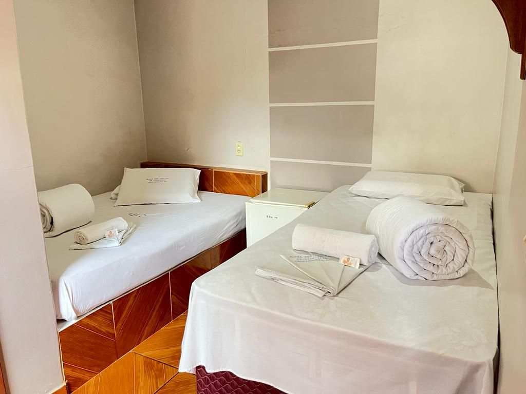 Quarto pequeno com duas camas de solteiro, lençóis brancos, toalhas dobradas e prateleiras embutidas na parede.