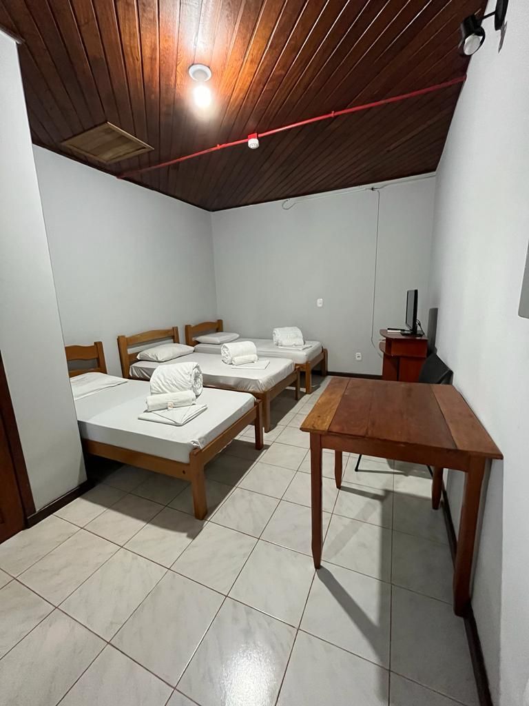 Quarto simples com duas camas de solteiro, teto de madeira, piso de azulejo e uma pequena mesa.