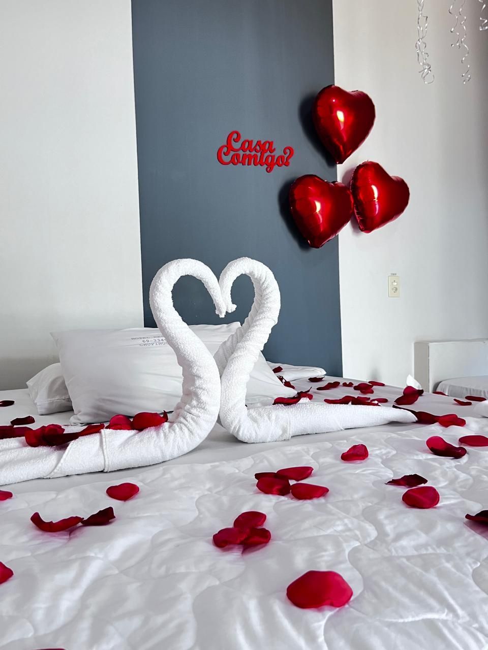 Quarto romântico com cisnes de toalha, pétalas de rosa vermelha e balões em forma de coração sobre uma cama branca.