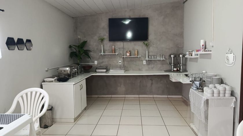 Cozinha cinza clara com piso de azulejo branco, TV fixada na parede, pia na bancada e cadeiras de plástico brancas em um ambiente iluminado.