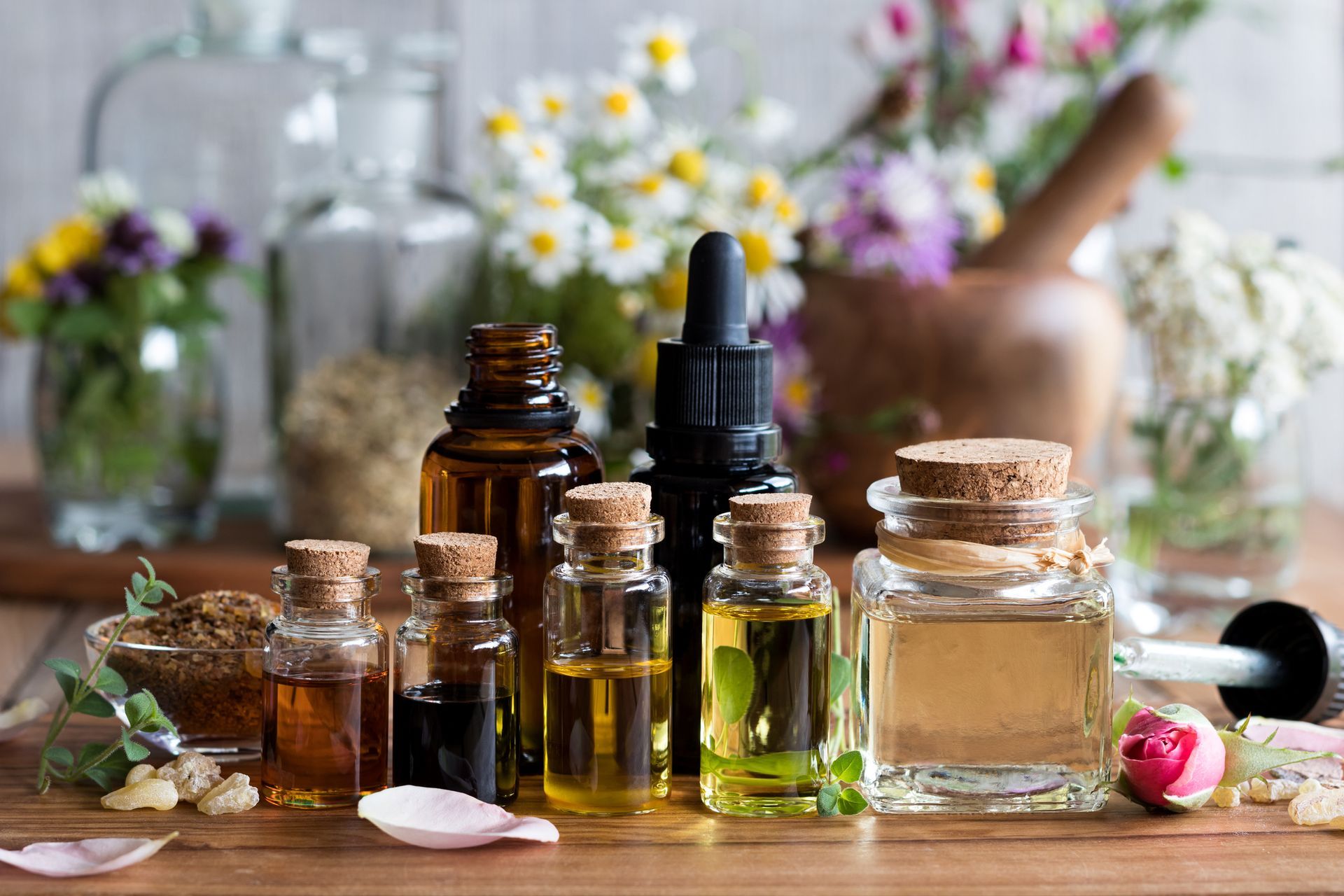 Bottles of essential oils with flowers, a mortar, and pestle on a wooden surface.