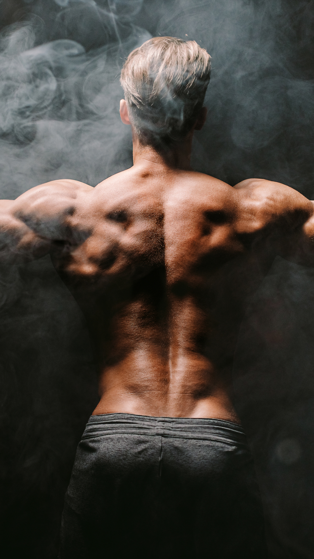 Muscular back of a person with blonde hair, arms outstretched, with smoke in background.