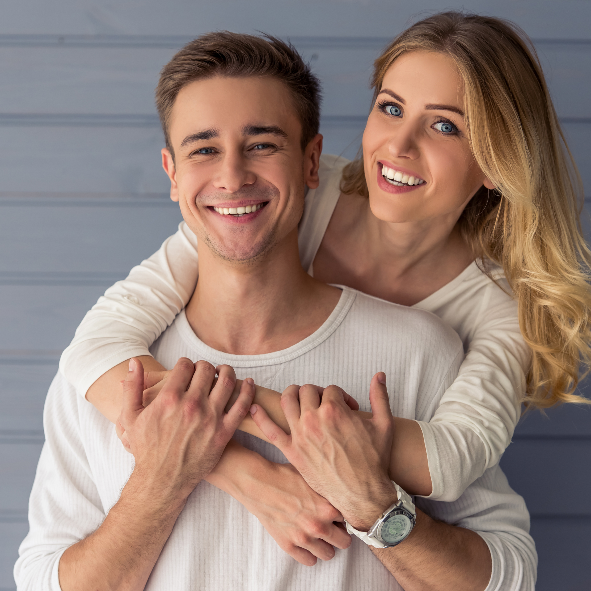 Smiling couple hugging, with the woman holding the man from behind against a light gray background