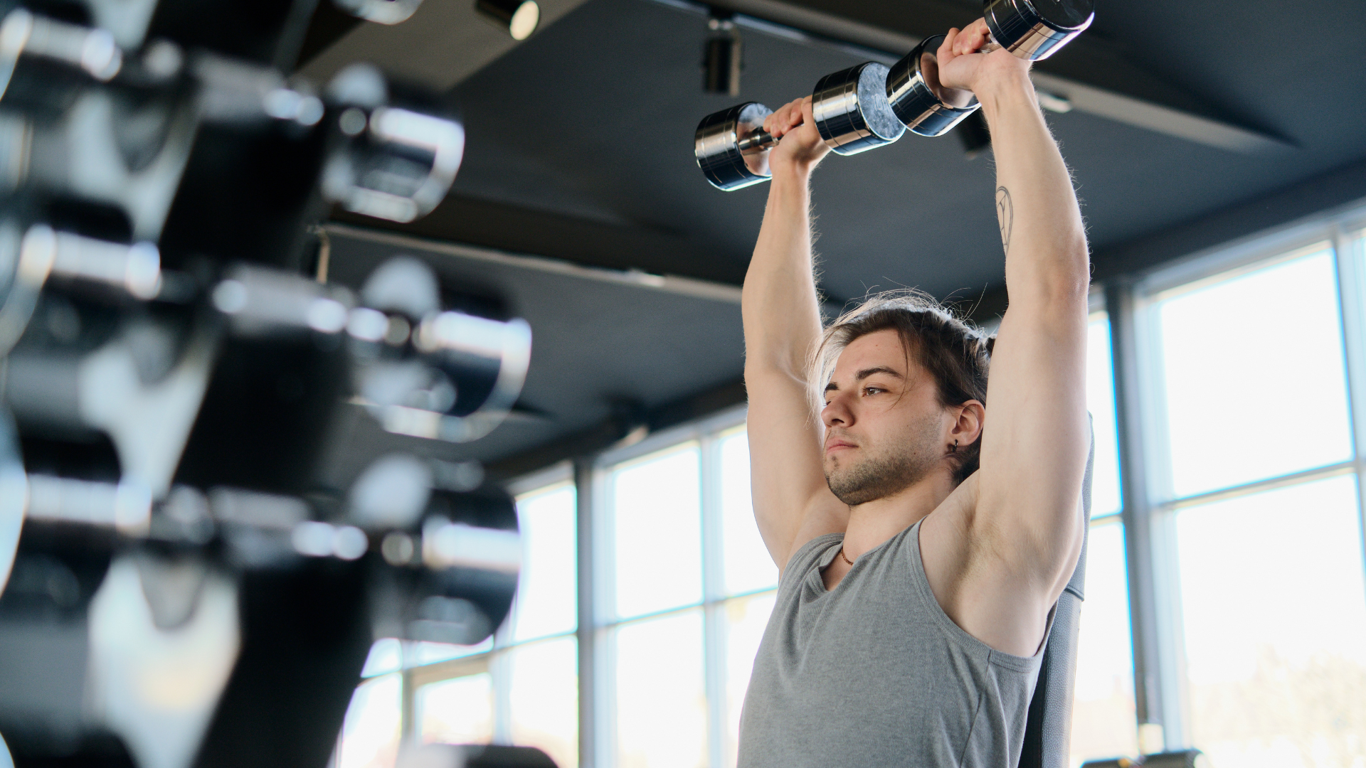 Man in gray tank top lifting dumbbells overhead in a gym.