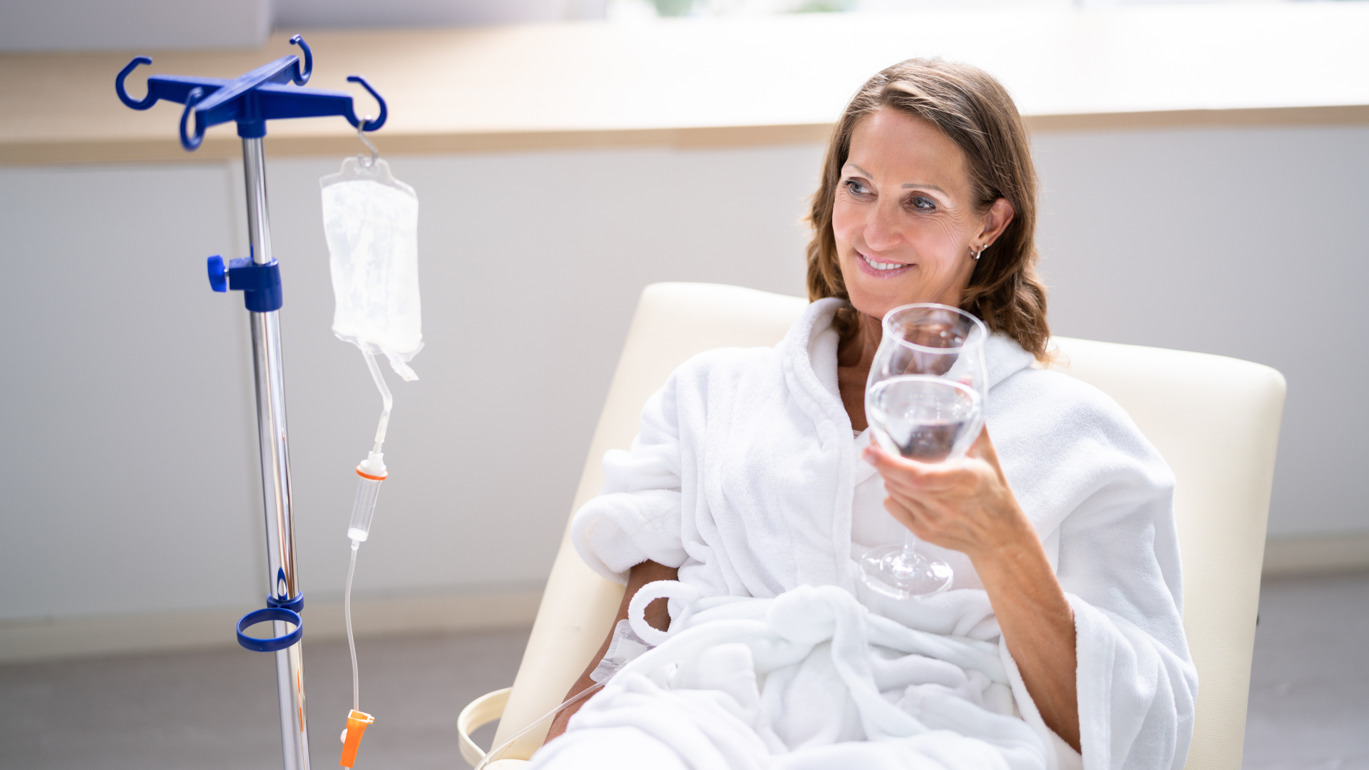Woman in bathrobe receiving IV, smiling, holding water glass; bright room with window.
