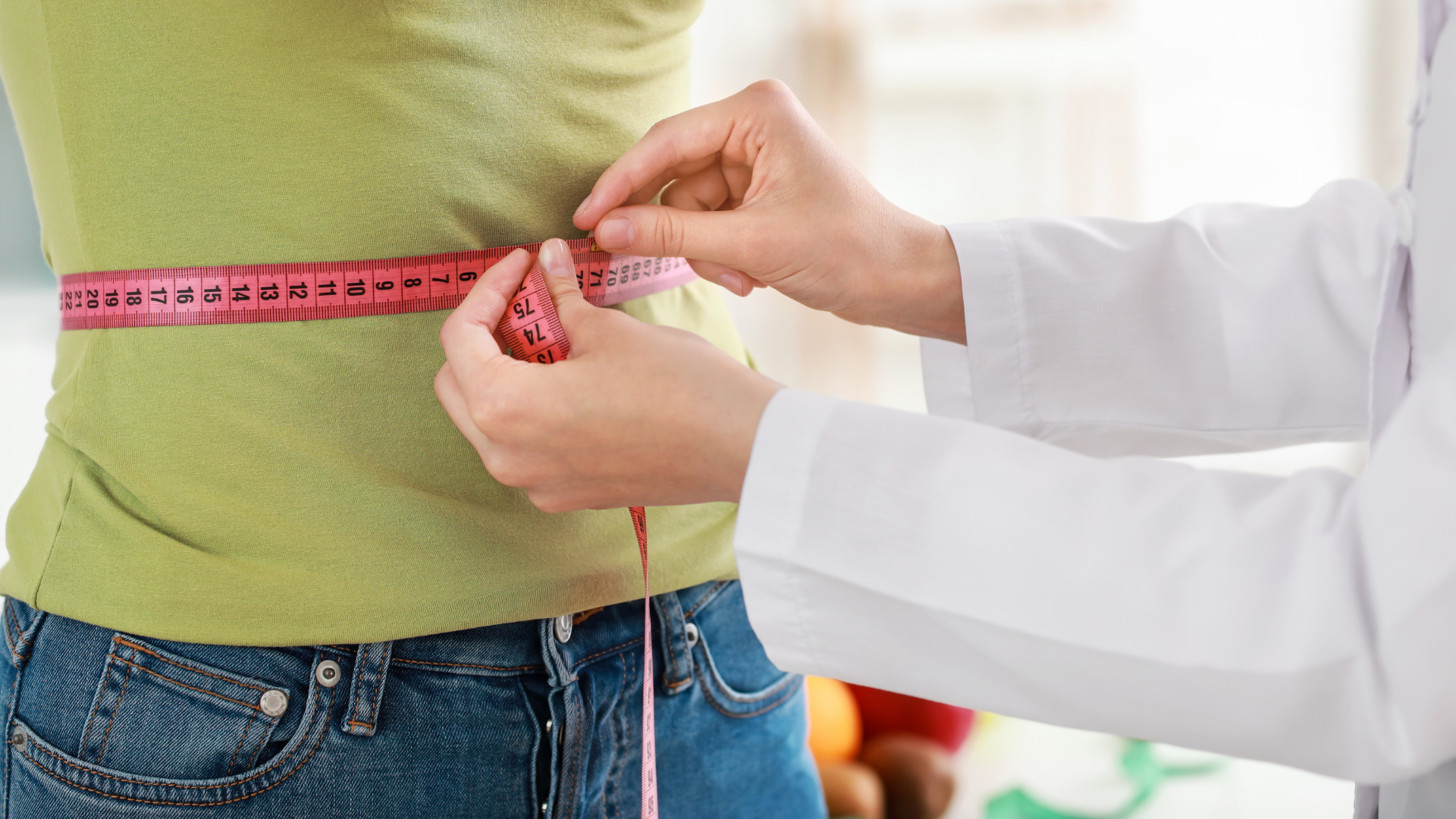 A person's waist is measured with a pink measuring tape by a person in a white coat.