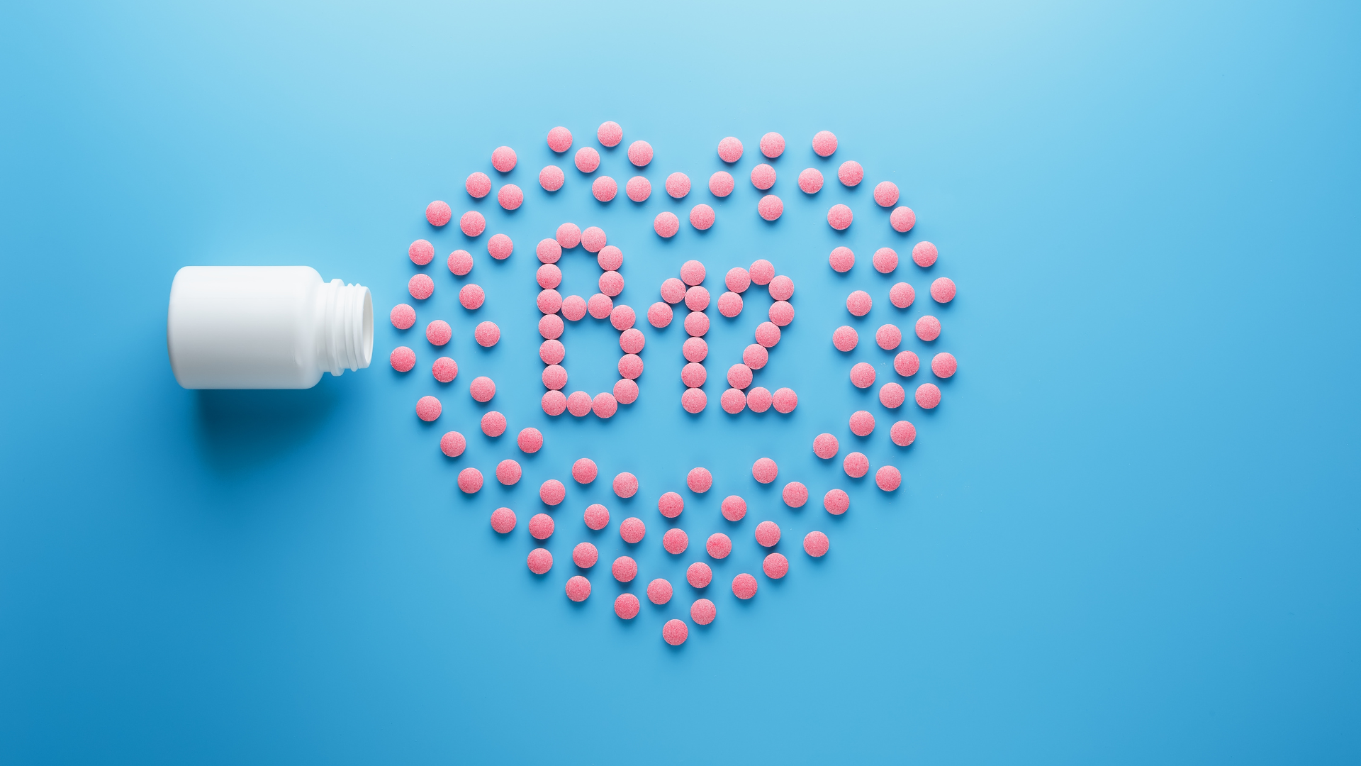 Pink B12 pills spilling from a white bottle in a heart shape on a blue background.