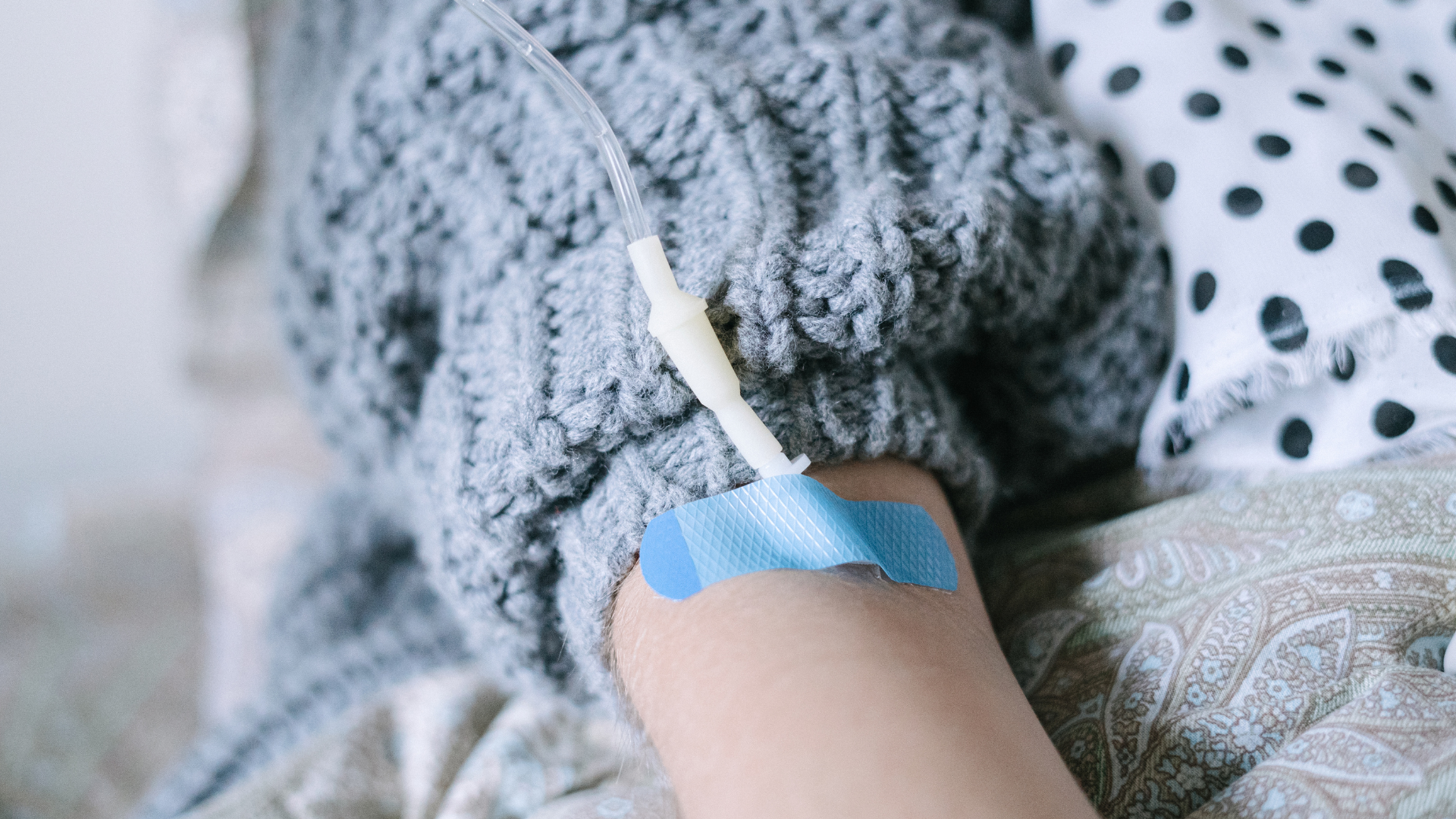 Woman in bathrobe receiving IV, smiling, holding water glass; bright room with window.
