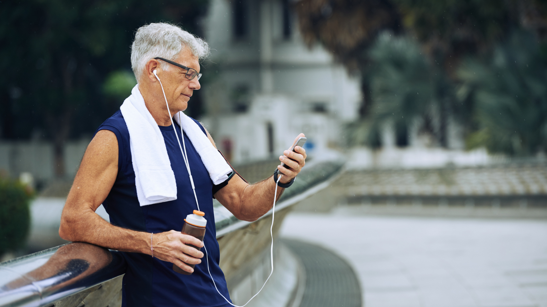 Older man in athletic wear checks phone, rests on railing, holding water bottle, wearing earbuds.