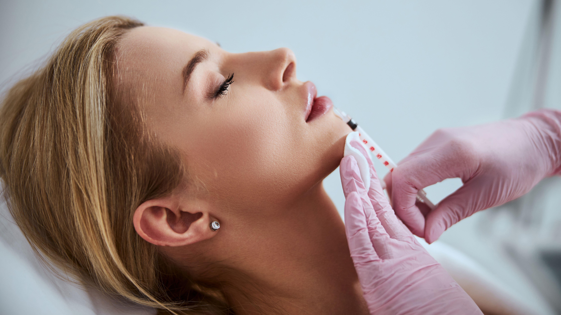 Woman receiving an injection in her jaw, hands in pink gloves, light setting.
