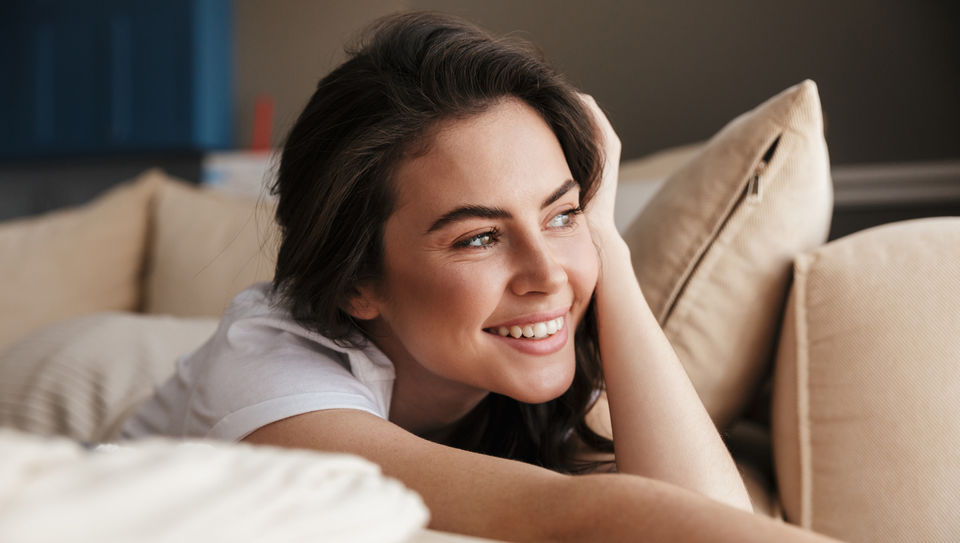 Woman smiles, reclining on a beige sofa, looking off-screen.