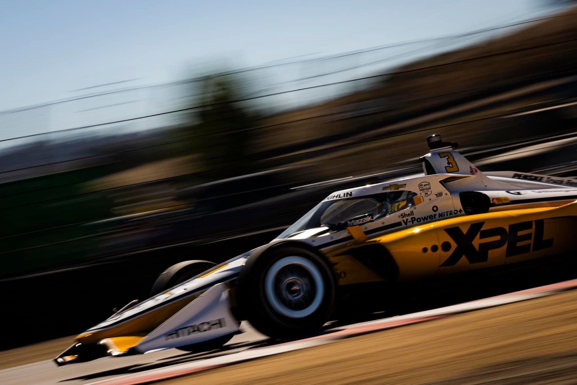 A yellow and black XPEL IndyCar race car speeding along a track, captured with motion blur.