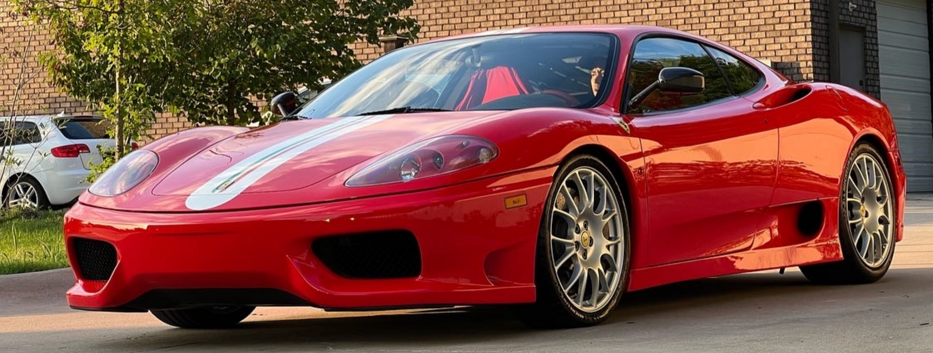 A red Ferrari 360 Modena with a white racing stripe parked on a driveway in front of a building.