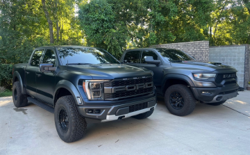 Two dark-colored pickup trucks, a Ford Raptor and a Ram TRX, parked side-by-side on a paved driveway.