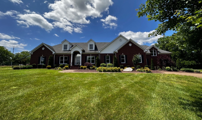 A single-story, red brick home with a white gable, dark roof, and two dormers, set behind a lush green lawn under blue sky.
